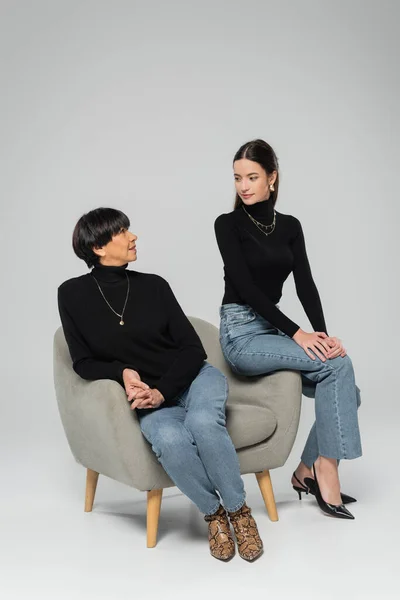 full length of asian mother and daughter in black turtlenecks and jeans posing on armchair and looking at each other on grey background