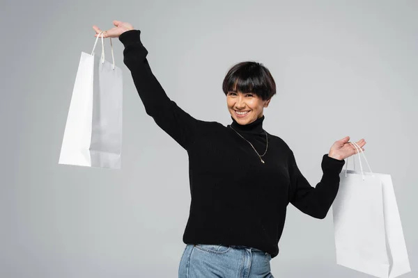 stock image cheerful asian woman in black turtleneck holding white shopping bags and smiling at camera isolated on grey