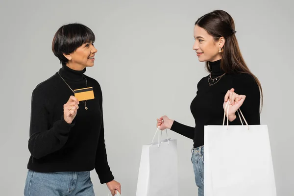 happy asian woman showing credit card to trendy daughter holding white shopping bags isolated on grey