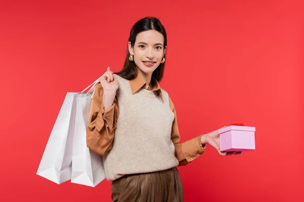 stock image young woman in stylish casual attire holding present and shopping bags while smiling at camera isolated on coral