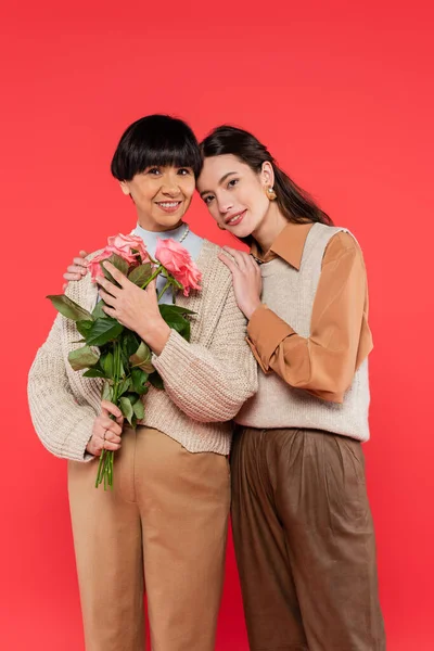 Stock image joyful asian daughter hugging smiling mother with bouquet of flowers isolated on coral 