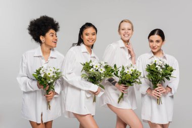 happy multicultural women in white shirts holding spring bouquets isolated on grey