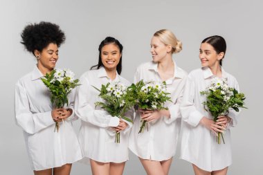 young interracial women in white shirts holding flowers and smiling isolated on grey clipart