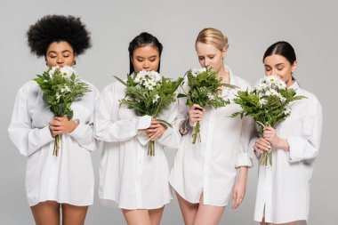  charming multiethnic women with closed eyes enjoying flavor of aromatic flowers isolated on grey