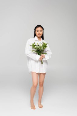 full length of asian woman with bare legs wearing white shirt and holding bouquet on grey background