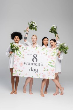 full length of cheerful multiethnic women holding flowers and greeting placard with womens day lettering on grey background clipart