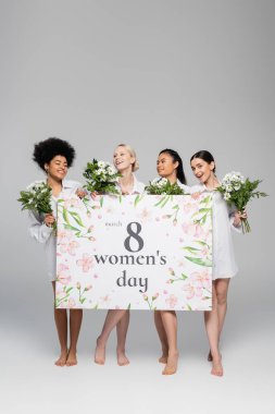 full length of barefoot multiethnic women holding flowers and placard with womens day lettering on grey background