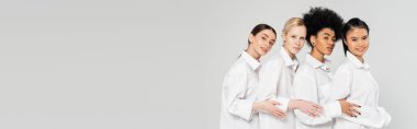 young multiethnic women wearing white shirts and looking at camera isolated on grey, banner
