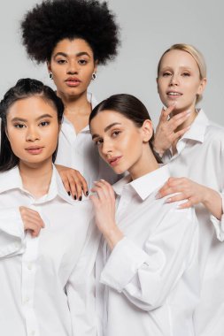 pretty multiethnic women wearing white shirts and looking at camera isolated on grey