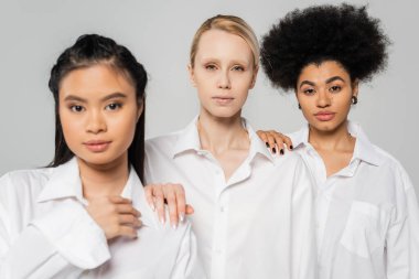 young blonde model looking at camera near asian and african american women in white shirts isolated on grey