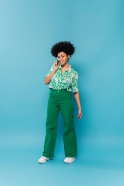 full length of happy african american woman in green pants and leaf print blouse talking on mobile phone on blue background