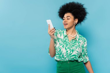 Yaprak desenli bluz giyen Afro-Amerikan kadın mavi üzerinde izole edilmiş cep telefonuna bakıyor.