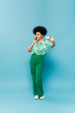 full length of smiling african american woman showing credit card while talking on smartphone on blue background