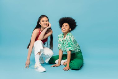 full length of cheerful african american and asian women in stylish casual attire posing on blue background