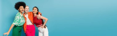 joyful multicultural women in trendy and colorful clothes embracing and smiling at camera isolated on blue, banner