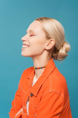 side view of joyful blonde woman in colorful beads and orange jacket isolated on blue
