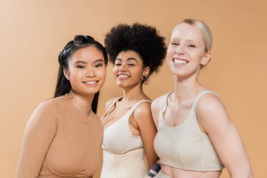 young and happy multiethnic women in underwear looking at camera isolated on beige