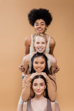 young and cheerful women in underwear posing and looking at camera isolated on beige