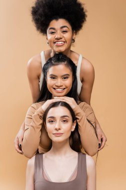 cheerful multicultural women in lingerie smiling at camera while posing isolated on beige