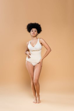 full length of curly african american woman in underwear posing with hand on hip on beige background