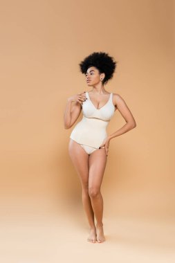 full length of barefoot african american woman in bodysuit posing with hand on hip and looking away on beige background clipart