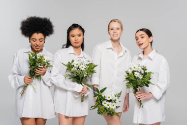 positive multicultural women in white shirts holding bouquets isolated on grey