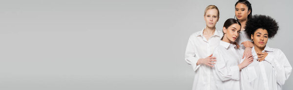 young multiethnic women in white shirts touching each other while posing isolated on grey, banner