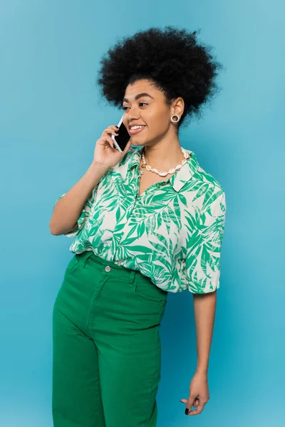 african american woman in green pants and trendy blouse smiling during conversation on smartphone isolated on blue