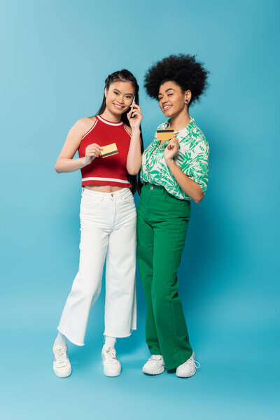 полная длина улыбающейся азиатской женщины, говорящей по мобильному телефону и показывающей кредитную карту рядом с африканским американским другом на синем фоне