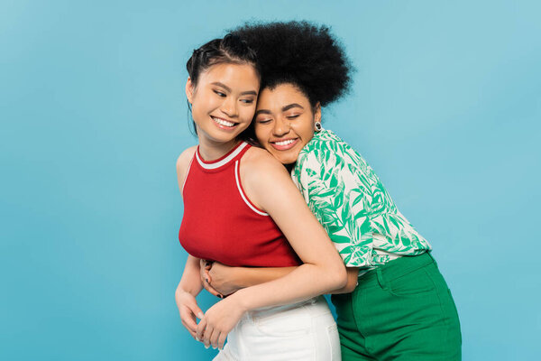 счастливая африканская американка с закрытыми глазами, обнимающая улыбающегося азиатского друга, изолированного на голубом