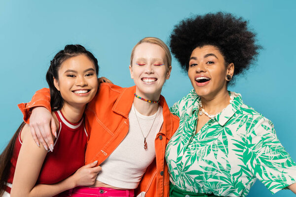 cheerful multiracial women in trendy colorful clothes embracing isolated on blue