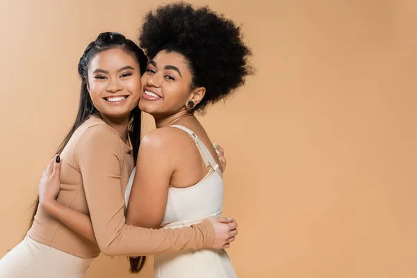 joyful asian and african american women in underwear embracing and looking at camera isolated on beige