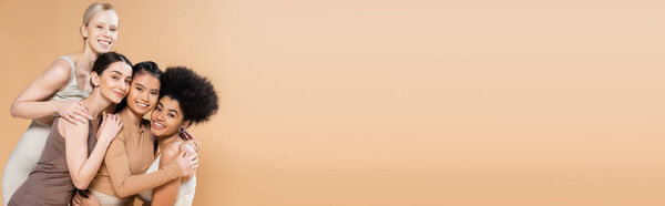 молодые и счастливые многоэтнические женщины в нижнем белье обнимающие и улыбающиеся в камеру изолированные на бежевом, баннере