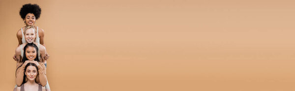 joyful multicultural women in underwear smiling at camera while posing isolated on beige, banner