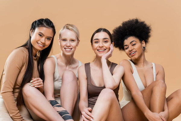 pleased multicultural models in underwear sitting and smiling at camera isolated on beige
