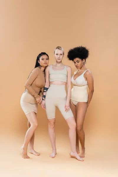 full length of young blonde woman in underwear looking at camera near brunette interracial models on beige background