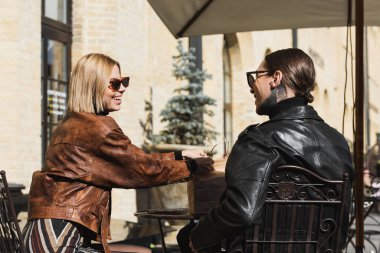 cheerful blonde woman in sunglasses holding paper bag while sitting on metallic bistro chair near tattooed boyfriend 