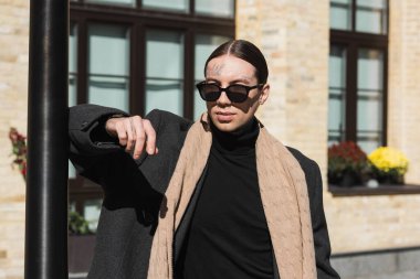 tattooed young man in coat and stylish sunglasses leaning on street pole 