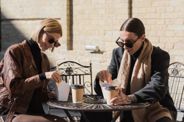 stylish couple eating asian food from carton boxes near paper cups on metallic bistro table 