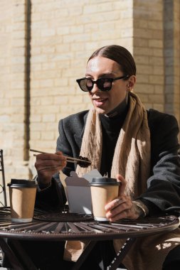 stylish tattooed man in sunglasses and coat holding chopsticks while eating asian food 