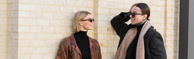 happy tattooed man flirting with happy woman in trendy sunglasses near brick wall, banner 