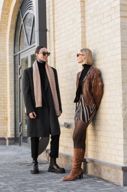 happy tattooed man talking with happy woman in trendy sunglasses near brick wall 