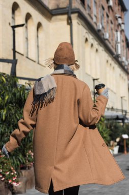 back view of blonde woman in beige coat and hat holding paper cup with coffee to go 
