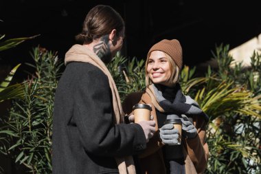 happy blonde woman in beige hat looking at tattooed boyfriend in coat while holding coffee to go 