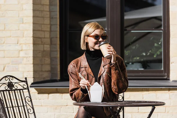 stock image happy blonde woman in sunglasses holding chopsticks near takeaway box with asian food and drinking coffee 