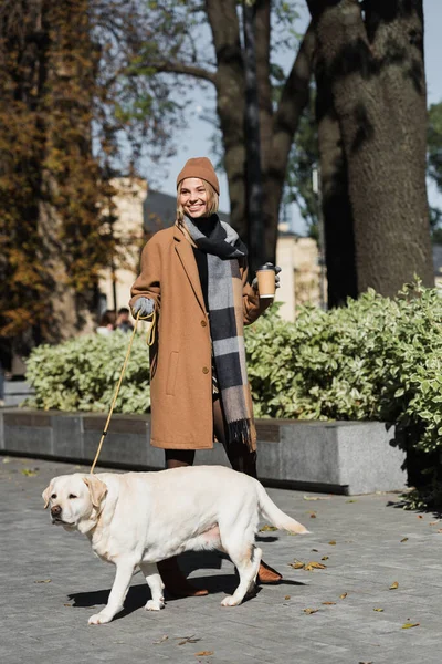 stock image full length of cheerful woman in hat and coat holding paper cup while walking with dog 