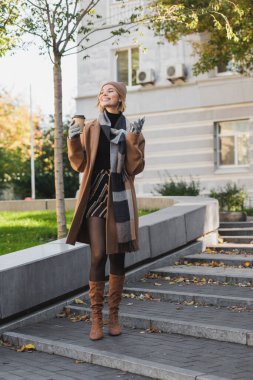 full length of happy blonde woman in hat and coat holding paper cup and smartphone outdoors 
