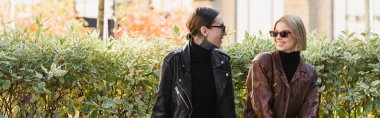 happy and stylish couple in sunglasses and leather jackets looking at each other outdoors during date, banner clipart