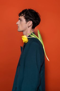 Side view of trendy brunette man with yellow tulip on shoulder on red background 