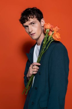 Portrait of stylish man in jacket holding roses near face on red background 